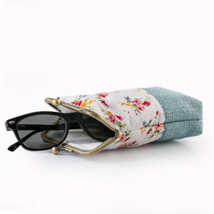 May include: Black sunglasses rest inside a floral print glasses case. The case features a white fabric with a pink and red floral pattern, a gold-colored metal frame, and a blue-green textured fabric on the sides.