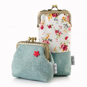 May include: Two vintage-style coin purses. One is a small, light blue purse with a red flower accent. The other is a tall, rectangular case with a floral pattern on a white background. Both have antique bronze clasps and the brand name "Shusha" on a tag.