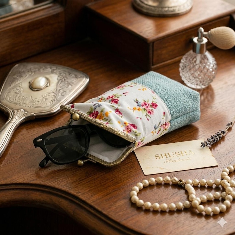 May include: A floral print glasses case with black sunglasses inside, resting on a wooden surface. The case has a white background with pink, yellow, and red flowers. A tag reads "SHUSHA Handmade". A silver hand mirror and perfume bottle are also visible.