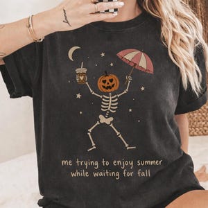 Op de afbeelding: Een antracietgrijs t-shirt met een afbeelding van een skelet met een pompoenhoofd dat een koffiebeker en een gestreepte paraplu vasthoudt onder een maansikkel en sterren. De tekst luidt: "me trying to enjoy summer while waiting for fall."