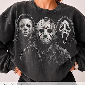 Op de afbeelding: Een antracietgrijs sweatshirt met ronde hals en een grafische print van drie horrorfilmfiguren. Het ontwerp toont een man met een bleek masker, een figuur met een ijshockeymasker en een spookachtig masker met een kap in zwart-wit.