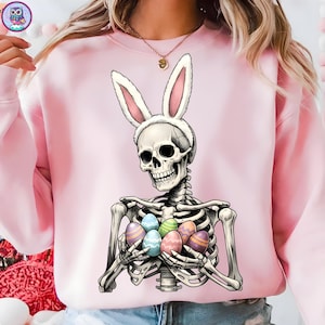 Puede incluir: Sudadera rosa claro con un esqueleto con orejas de conejo y huevos de Pascua coloridos. El diseño gráfico combina el tema de la Pascua con un tema espeluznante. Los huevos son multicolores.