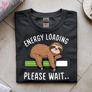 Puede incluir: Camiseta negra con un diseño de perezoso. El perezoso está acostado sobre una barra de carga verde y blanca, con el texto "ENERGY LOADING PLEASE WAIT.." en blanco arriba y abajo.