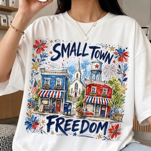 May include: White t-shirt with a colourful graphic depicting a small town scene. The design includes buildings, a water tower, fireworks, and the words "SMALL TOWN FREEDOM" in blue and red.