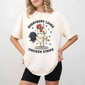 May include: Cream-colored t-shirt featuring a cartoon rooster wearing sunglasses and holding onto a pole. The text "EVERYBODY LOVES CHICKEN STRIPS" is printed above and below the rooster graphic. The shirt is a casual, unisex style.