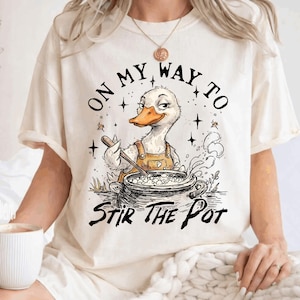 May include: Cream-colored t-shirt with a graphic of a duck wearing an apron, stirring a pot. Text reads "ON MY WAY TO STIR THE POT" in black. The duck has a smug expression. Stars and steam are also in the design.