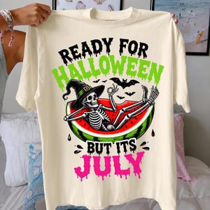 May include: A cream-colored t-shirt featuring a graphic of a skeleton wearing a witch hat, lounging inside a watermelon slice. The text reads "READY FOR HALLOWEEN" in green and "BUT ITS JULY" in pink, with black bats surrounding the skeleton.