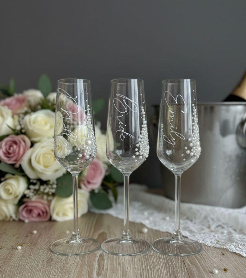 Puede incluir: Tres copas de champ&aacute;n de cristal transparente con tallos delgados y bases circulares. Cada copa presenta letras blancas en cursiva y un grupo de peque&ntilde;as perlas blancas adheridas al lateral. El texto en las copas dice "Bride" y "Emily".