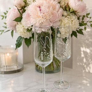 Puede incluir: Dos copas de champ&aacute;n de cristal transparente con texto en blanco y adornos de perlas. Una copa tiene grabado el nombre Sam y la palabra Groom, mientras que la otra tiene el nombre Amy. Ambas copas cuentan con un tallo alto y fino.