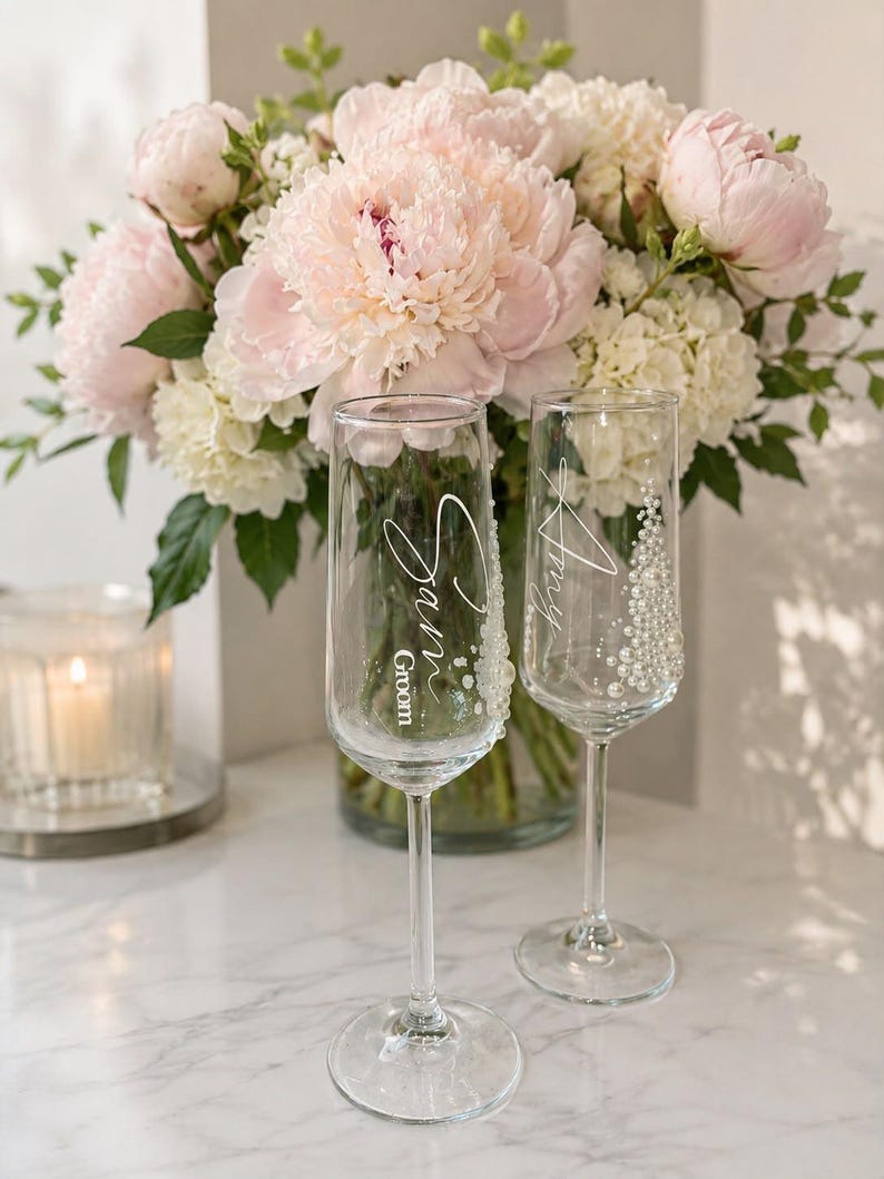 Puede incluir: Dos copas de champ&aacute;n de cristal transparente con texto en blanco y adornos de perlas. Una copa tiene grabado el nombre Sam y la palabra Groom, mientras que la otra tiene el nombre Amy. Ambas copas cuentan con un tallo alto y fino.