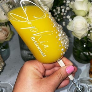 Puede incluir: Una copa de champ&aacute;n de cristal transparente llena de l&iacute;quido amarillo, con el texto en letra blanca Sophia y la palabra Bridesmaid debajo. El lateral de la copa est&aacute; decorado con una hilera vertical de perlas blancas de varios tama&ntilde;os.