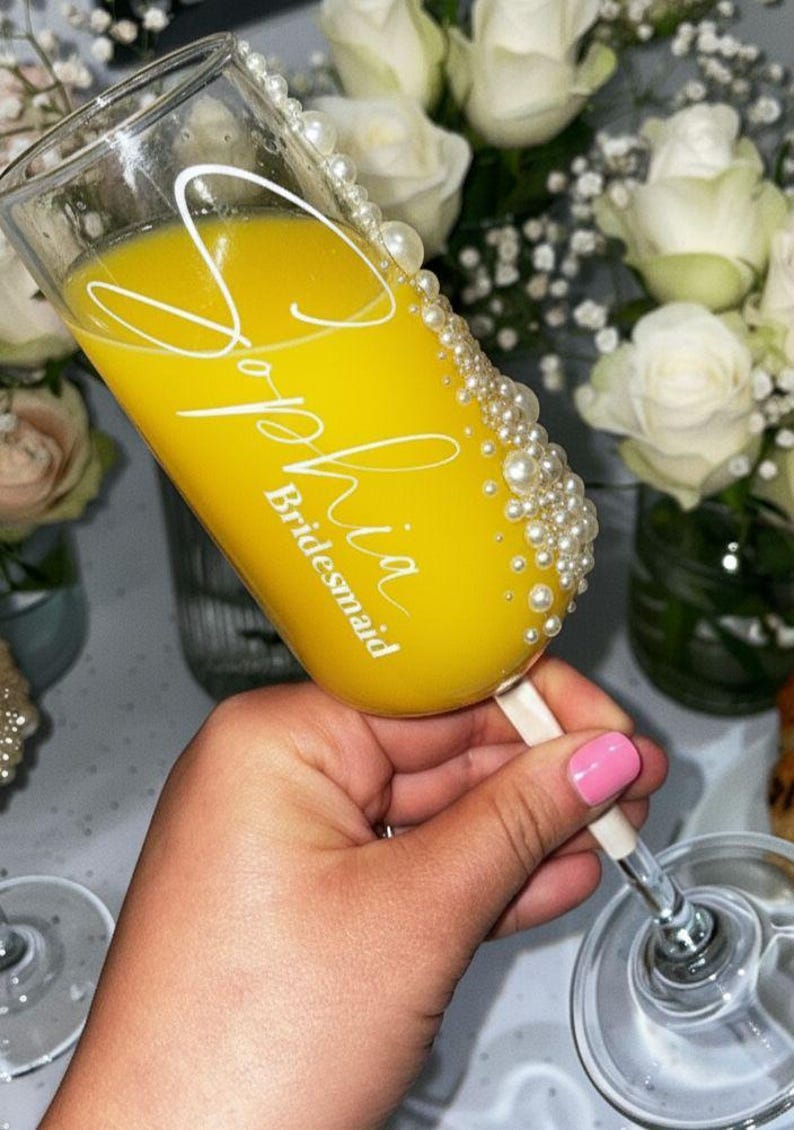 Puede incluir: Una copa de champ&aacute;n de cristal transparente llena de l&iacute;quido amarillo, con el texto en letra blanca Sophia y la palabra Bridesmaid debajo. El lateral de la copa est&aacute; decorado con una hilera vertical de perlas blancas de varios tama&ntilde;os.