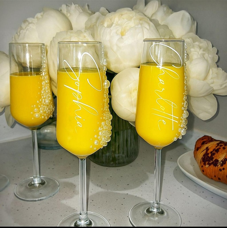 Puede incluir: Tres copas de champ&aacute;n de cristal transparente llenas de un l&iacute;quido amarillo brillante. Dos copas tienen texto cursivo blanco que dice Sophia y Charlotte. Cada copa est&aacute; decorada con un racimo vertical de perlas blancas en un lado.
