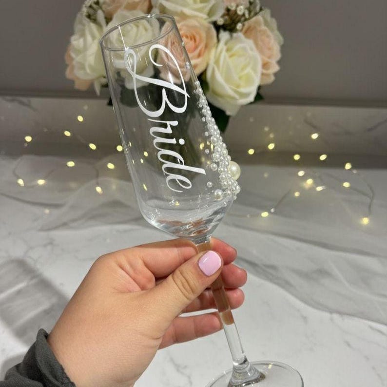 Puede incluir: Una copa de champ&aacute;n de cristal transparente con la palabra Bride escrita en letra cursiva blanca. El lateral de la copa est&aacute; decorado con una disposici&oacute;n vertical de peque&ntilde;as perlas blancas de varios tama&ntilde;os, desde el borde hasta el tallo.