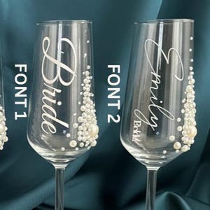 Puede incluir: Dos copas de champ&aacute;n de cristal transparente con texto blanco y adornos de perlas. La copa izquierda muestra la palabra Bride con el texto FONT 1, mientras que la derecha muestra el nombre Emily con el texto FONT 2 y la palabra Bride debajo.