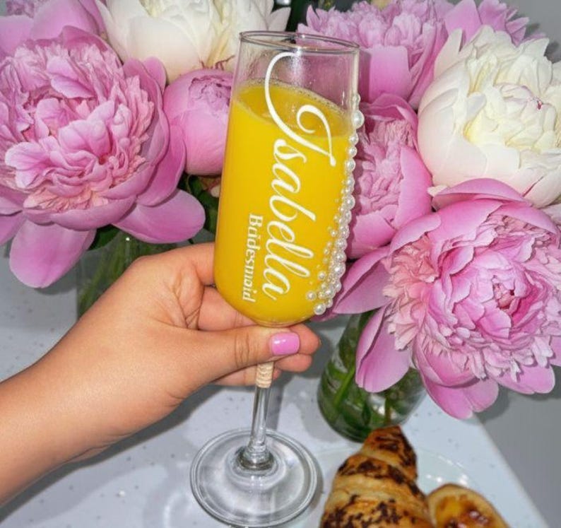 Puede incluir: Una copa de champ&aacute;n de cristal transparente llena de zumo de naranja, con el texto blanco Isabella y Bridesmaid impreso verticalmente. El lateral de la copa est&aacute; decorado con una fila vertical de peque&ntilde;as perlas blancas a lo largo del borde y el tallo.