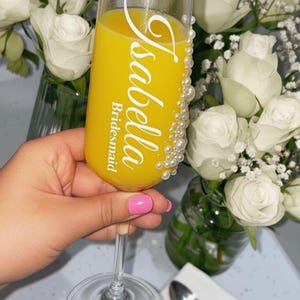 Puede incluir: Una copa de champ&aacute;n de cristal transparente llena de zumo de naranja, con el texto blanco Isabella y Bridesmaid impreso en el frente, y un grupo vertical de peque&ntilde;as perlas blancas falsas adheridas al lado de la copa.