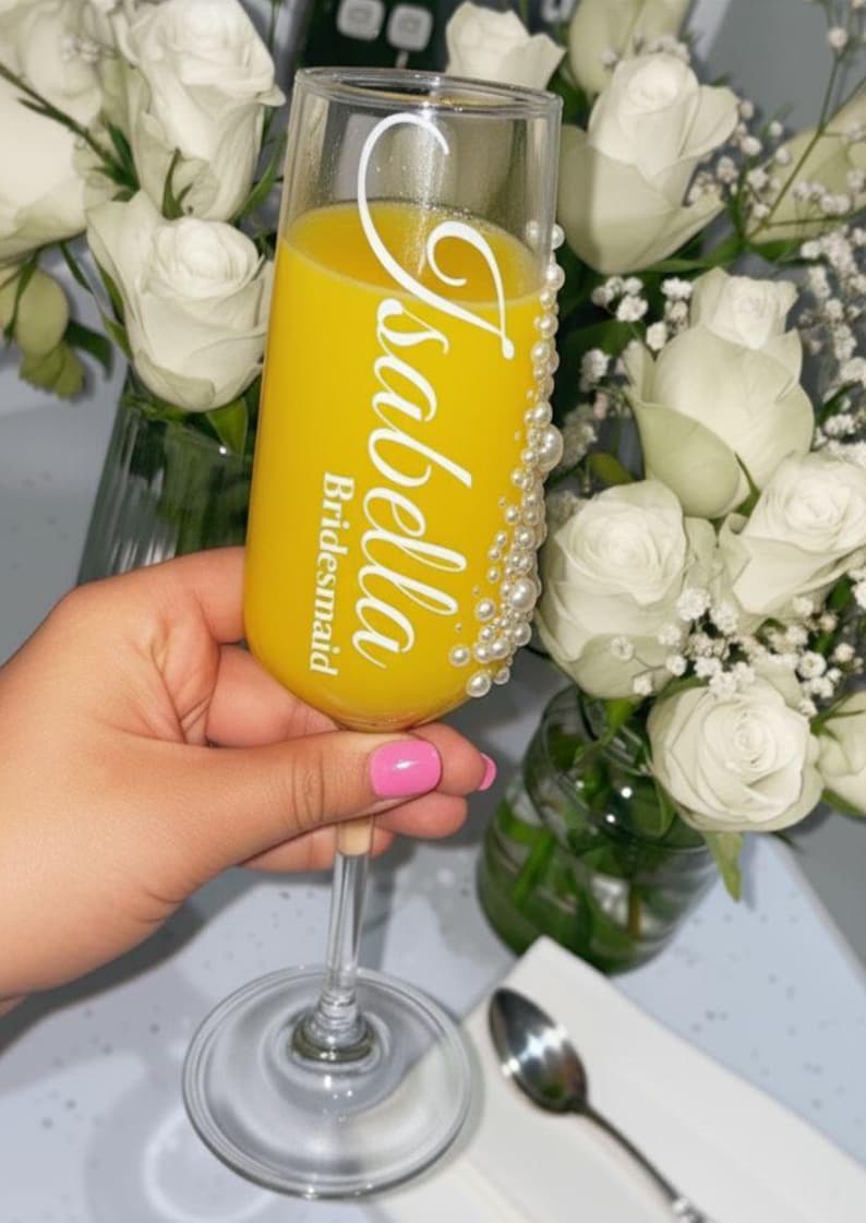 Puede incluir: Una copa de champ&aacute;n de cristal transparente llena de zumo de naranja, con el texto blanco Isabella y Bridesmaid impreso en el frente, y un grupo vertical de peque&ntilde;as perlas blancas falsas adheridas al lado de la copa.