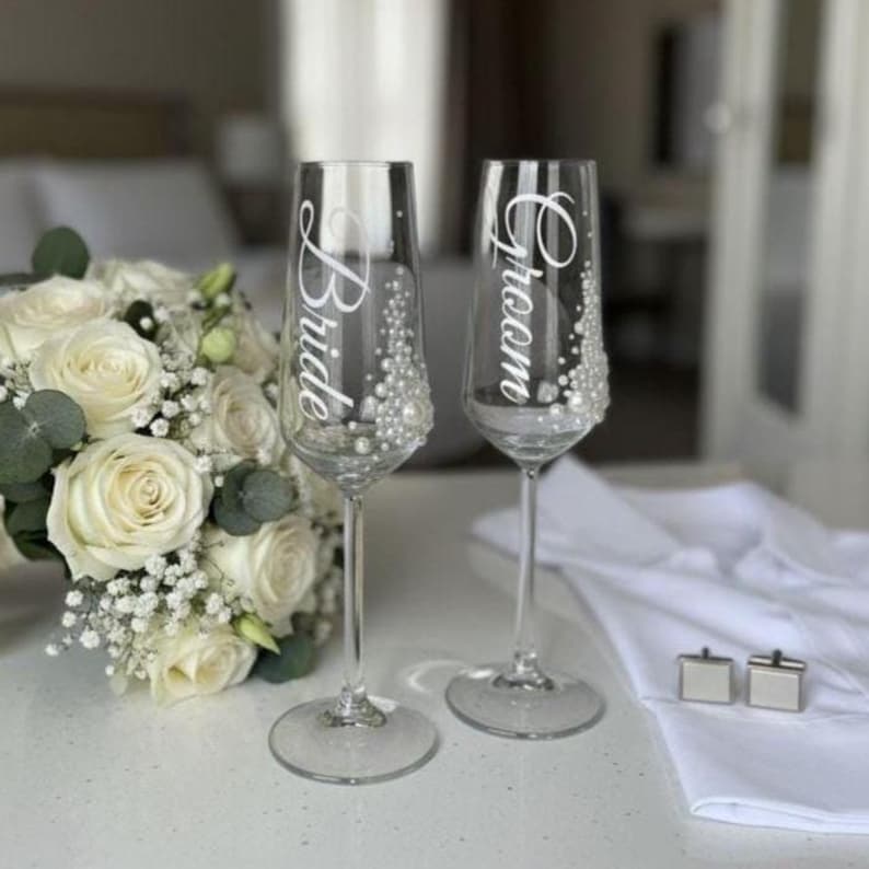 Puede incluir: Dos copas de champ&aacute;n de cristal transparente con texto en letra cursiva blanca que dice Bride y Groom, decoradas con peque&ntilde;as perlas blancas a lo largo del lado de cada cuenco. Las copas est&aacute;n junto a un ramo de rosas blancas y tela blanca.