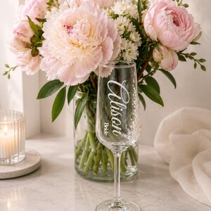 Puede incluir: Una copa de champ&aacute;n de cristal transparente con el texto Alison y Bride impreso en blanco en letra cursiva. La copa est&aacute; colocada frente a un jarr&oacute;n de cristal lleno de un ramo de peon&iacute;as de color rosa claro y peque&ntilde;as flores blancas.
