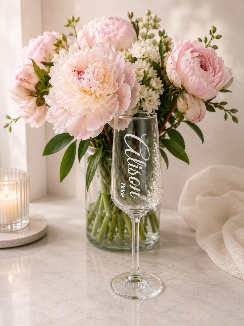 Puede incluir: Una copa de champ&aacute;n de cristal transparente con el texto Alison y Bride impreso en blanco en letra cursiva. La copa est&aacute; colocada frente a un jarr&oacute;n de cristal lleno de un ramo de peon&iacute;as de color rosa claro y peque&ntilde;as flores blancas.