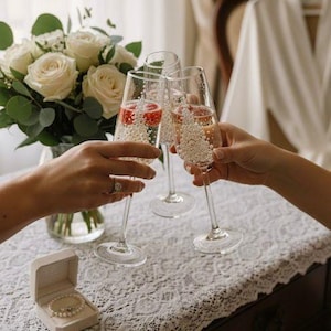 Puede incluir: Tres copas de champ&aacute;n de cristal transparente llenas de una bebida espumosa y peque&ntilde;as perlas decorativas blancas, situadas junto a un ramo de rosas blancas y un peque&ntilde;o joyero abierto con una pulsera de perlas sobre un mantel de encaje.