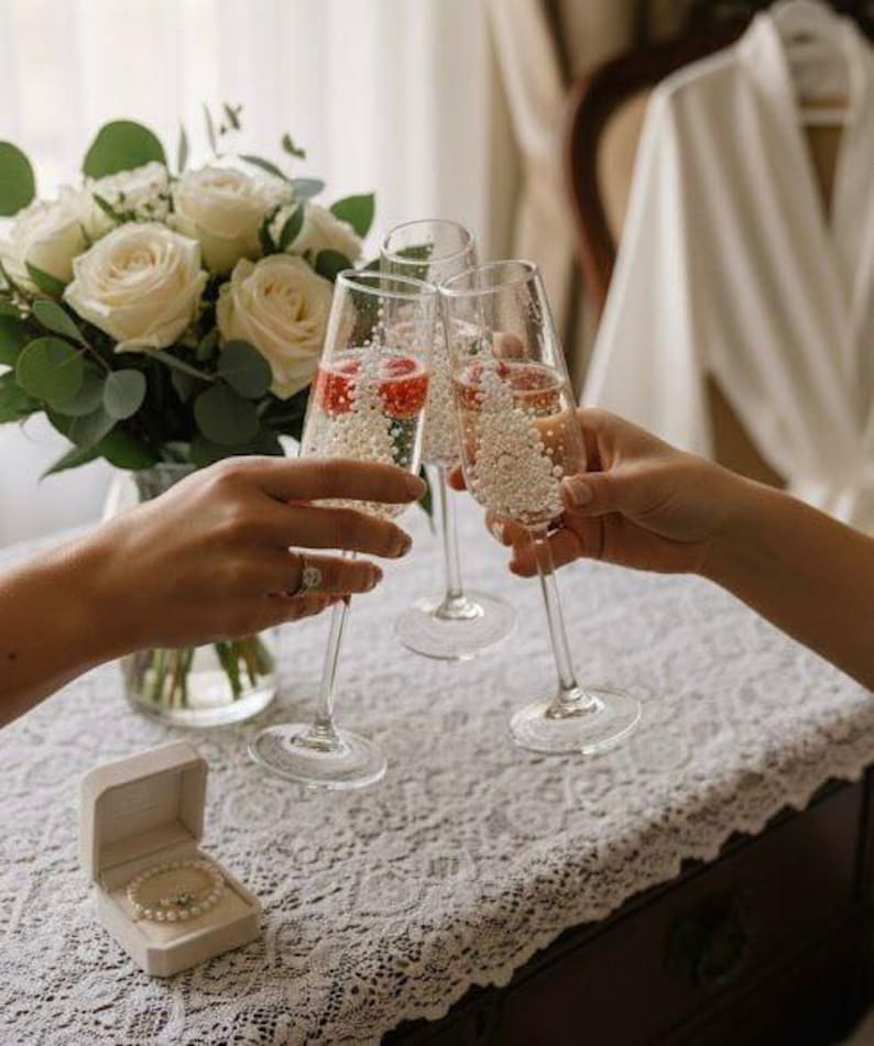 Puede incluir: Tres copas de champ&aacute;n de cristal transparente llenas de una bebida espumosa y peque&ntilde;as perlas decorativas blancas, situadas junto a un ramo de rosas blancas y un peque&ntilde;o joyero abierto con una pulsera de perlas sobre un mantel de encaje.