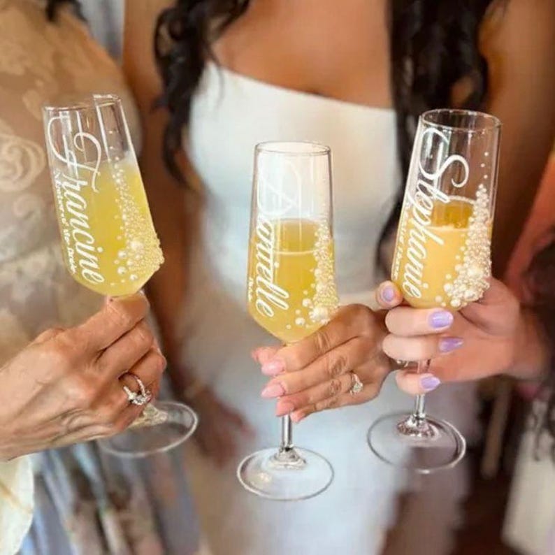Puede incluir: Tres copas de champ&aacute;n de cristal transparente llenas de un l&iacute;quido amarillo. Cada copa presenta letras cursivas blancas que dicen Francine, Danielle o Stephanie, y est&aacute; decorada con peque&ntilde;as cuentas blancas tipo perla en vertical.