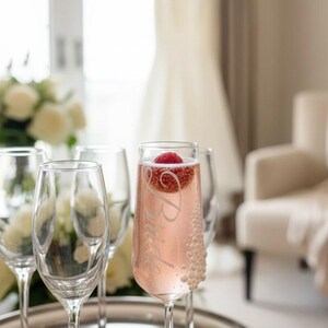 Puede incluir: Una copa de champ&aacute;n de cristal transparente llena de una bebida espumosa rosa, con la palabra Bride grabada en cursiva, una frambuesa flotando en la parte superior y peque&ntilde;os detalles en forma de perlas blancas en el lateral.