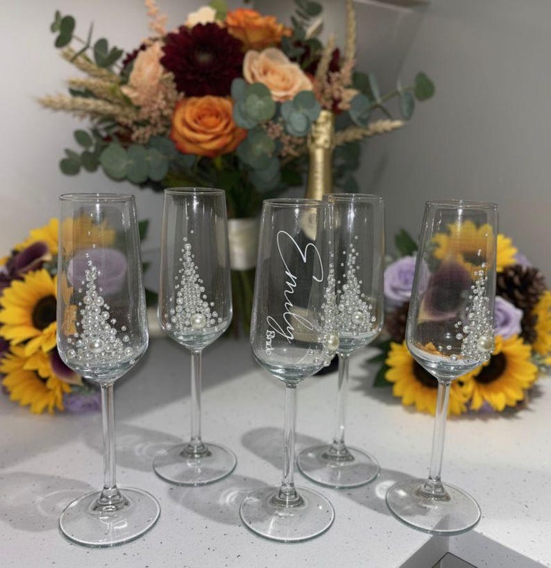 Puede incluir: Cinco copas de champ&aacute;n de cristal transparente con tallos delgados y bases circulares. Cuatro copas presentan un dise&ntilde;o triangular decorativo de peque&ntilde;as perlas blancas y cristales. Una copa central est&aacute; personalizada con el texto Emily Bride en blanco.