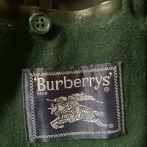 Könnte beinhalten: Nahaufnahme eines dunkelgrünen Kleidungsstücks mit Knopf und Etikett. Das Etikett, mit einer genähten Umrandung, zeigt "Burberrys" in weißem Text und ein Ritteremblem.