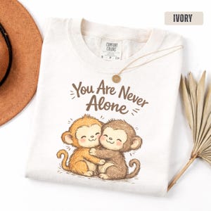 Puede incluir: Camiseta de color marfil con el texto "You Are Never Alone" y un gráfico de dos monos abrazándose. La camiseta tiene cuello redondo y mangas cortas. Un collar dorado con un colgante está sobre la camiseta.