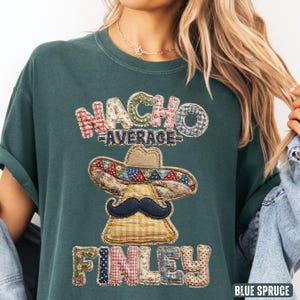 Puede incluir: Camiseta de color verde azulado con el texto "NACHO AVERAGE FINLEY" en un diseño de patchwork. El diseño incluye un sombrero y un bigote. La camiseta es de corte holgado.