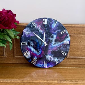 May include: Round wall clock with a swirling galaxy design in shades of purple, blue, and white. The clock face features silver Roman numerals and hands. The clock is displayed on a wooden surface.