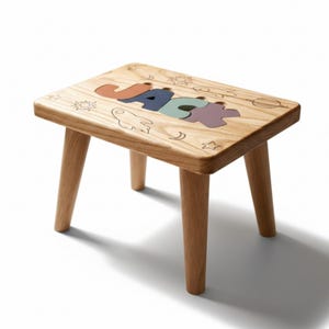 Puede incluir: Un taburete de madera con el nombre "JACK" escrito en letras coloridas. El taburete tiene cuatro patas de madera y está decorado con ilustraciones temáticas del espacio. El taburete es de madera clara.