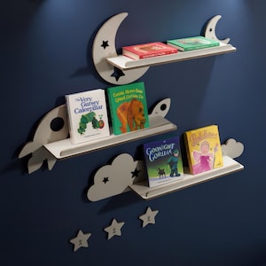 May include: A set of three decorative shelves in the shape of a moon, rocket, and cloud, holding children's books. The shelves are white with gray accents and star cutouts, mounted on a dark blue wall. Books include "The Very Gutsy Caterpillar" and "Pinkalicious".