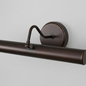 May include: A bronze-coloured picture light. The light has a cylindrical housing, a curved arm, and a circular wall mount. The finish is a dark, metallic brown. Designed to illuminate artwork, it is suitable for galleries.
