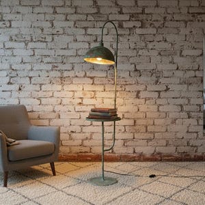 May include: A vintage-style floor lamp with a green patina finish, with a small side table holding books. The lamp lights a room with a grey armchair and a patterned rug. A white brick wall forms the backdrop.