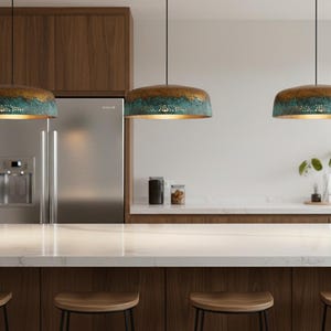 May include: A modern kitchen with three pendant lights. The lights have a bronze top, turquoise middle, and gold interior. The kitchen features a white countertop, wooden cabinets, and a stainless steel refrigerator. Wooden stools are at the island.