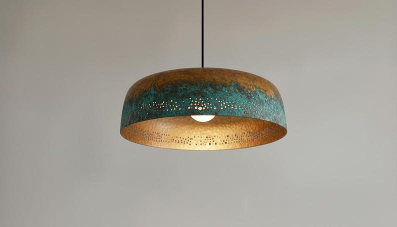 May include: A pendant light fixture with a domed, metallic shade. The shade has a bronze-colored top that transitions to a turquoise-green hue. The bottom of the shade is gold-colored and perforated with small holes. A single light bulb is visible.