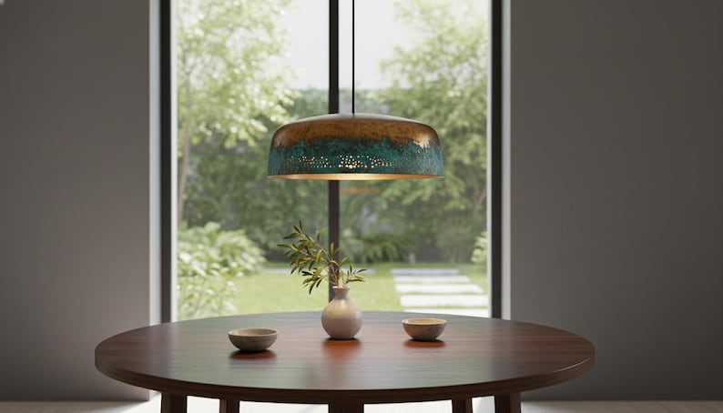 May include: A bronze and teal pendant light hangs above a dark wood table. The light fixture has a textured finish and a perforated lower section. A small white vase with greenery and two small bowls are on the table.