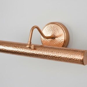 May include: A copper-colored picture light with a hammered texture. The light has a cylindrical shade, a curved arm, and a circular wall mount. The fixture is designed to illuminate artwork.