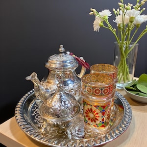 Könnte beinhalten: Ein silbernes Teeservice mit aufwendigen Details, darunter eine Teekanne, eine Zuckerdose und ein Serviertablett. Zwei dekorative Gläser mit farbenfrohen geometrischen Mustern stehen auf dem Tablett. Eine Vase mit weißen Blumen und eine kleine Schale mit grünen Blättern vervollständigen das Arrangement.