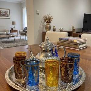 Può includere: Un servizio da tè in argento con un design decorativo, tra cui una teiera e sei bicchieri colorati su un vassoio d'argento. I bicchieri sono nelle tonalità di blu, giallo e marrone. Il set è su un tavolo di legno.