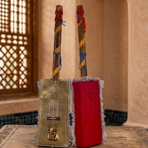 Peut inclure: Deux instruments de musique à cordes avec des corps rectangulaires. Un corps est en métal doré, l'autre est recouvert de tissu rouge. Les deux ont de longs manches en bois rayés jaune et marron, ornés de liens rouges et bleus et de franges argentées.