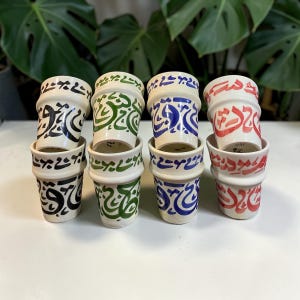 Puede incluir: Ocho pequeñas tazas de cerámica blanca con caligrafía árabe en negro, verde, azul y rojo. Las tazas están apiladas por parejas. El diseño se repite en cada taza. La disposición muestra los intrincados patrones y colores.