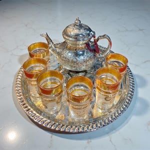 Puede incluir: Un juego de té plateado con una tetera decorativa y seis vasos con bordes dorados, dispuestos sobre una bandeja plateada. La tetera tiene patrones intrincados y una borla. Los vasos son transparentes con detalles dorados.