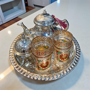 Puede incluir: Un juego de té plateado sobre una bandeja redonda. El juego incluye una tetera, un azucarero y dos vasos decorativos. Los vasos tienen patrones florales coloridos. Una borla cuelga de la tetera.