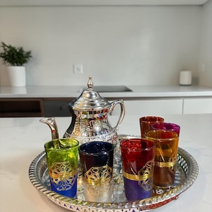 Può includere: Un servizio da tè in argento con una teiera e un vassoio con sei bicchieri colorati. I bicchieri sono nelle tonalità di verde, blu, rosso, arancione e viola, con disegni in filigrana dorata. Il set è su un piano di lavoro bianco.