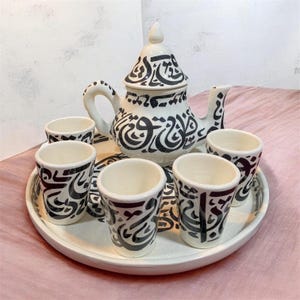 May include: A white ceramic tea set with black calligraphy-style designs. The set includes a teapot with a lid, six small cups, and a round serving tray. The tea set is on a pink surface.